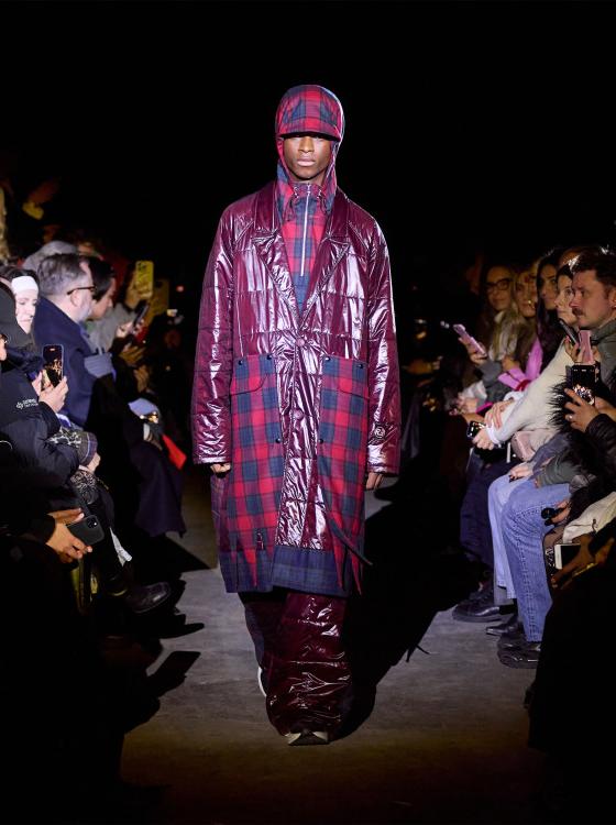 A man wearing a glossy burgundy Rolf Ekroth puffer coat over a red tartan check ensemble with a matching flat cap