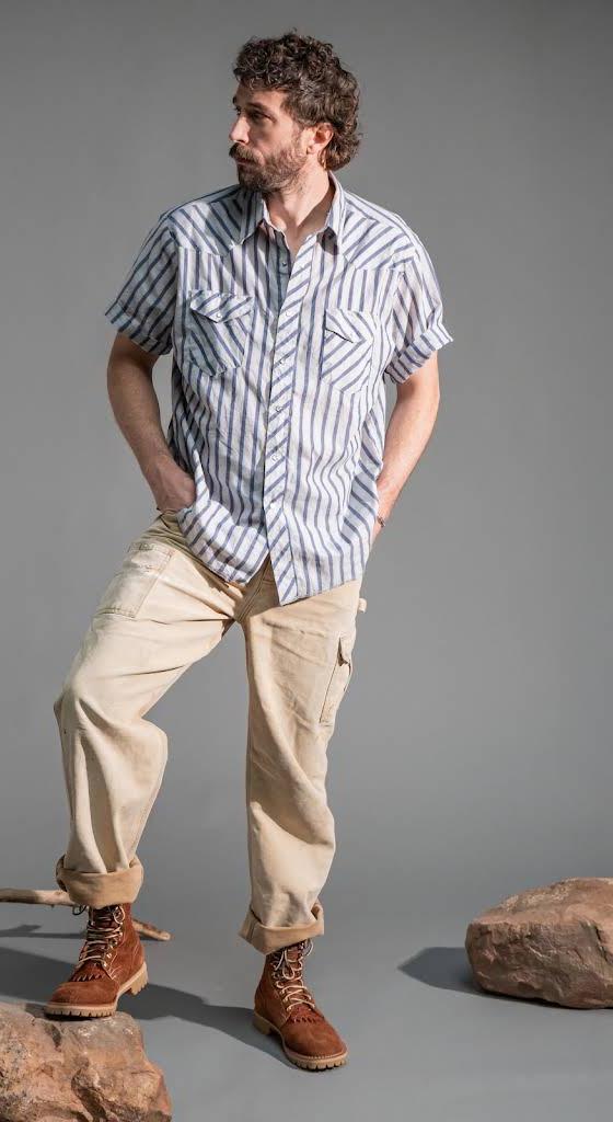A man wearing a blue striped short-sleeve shirt, cream cargo trousers and tan leather Rose Anvil boots