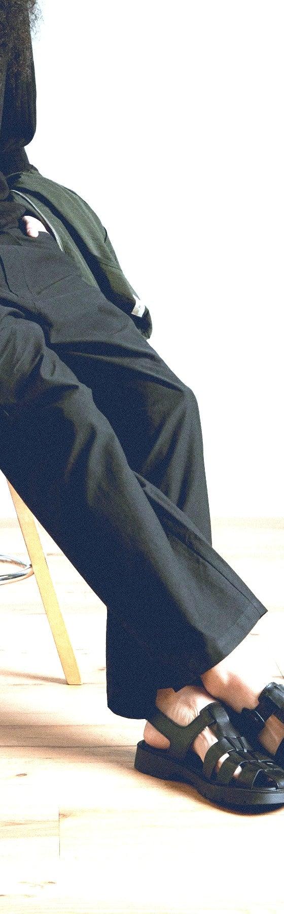 A person seated on a wooden stool wearing dark trousers, black socks and black sandals