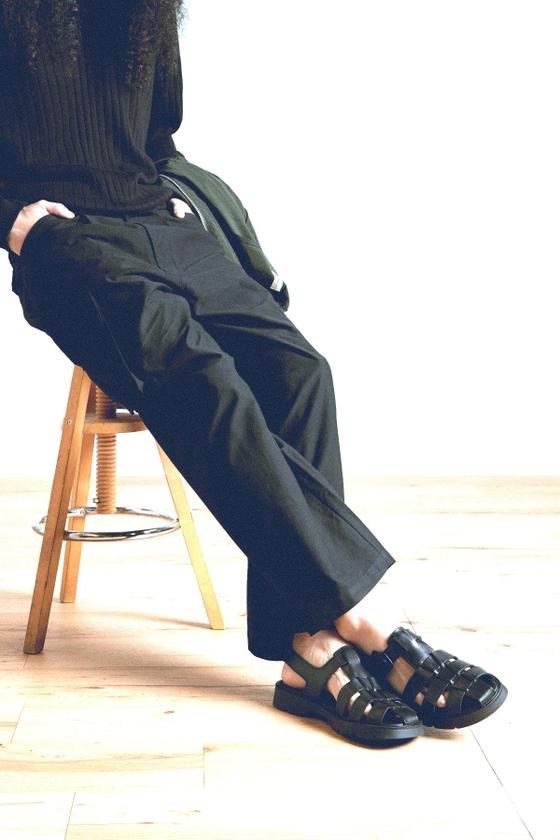 A person seated on a wooden stool wearing dark trousers, black socks and black sandals
