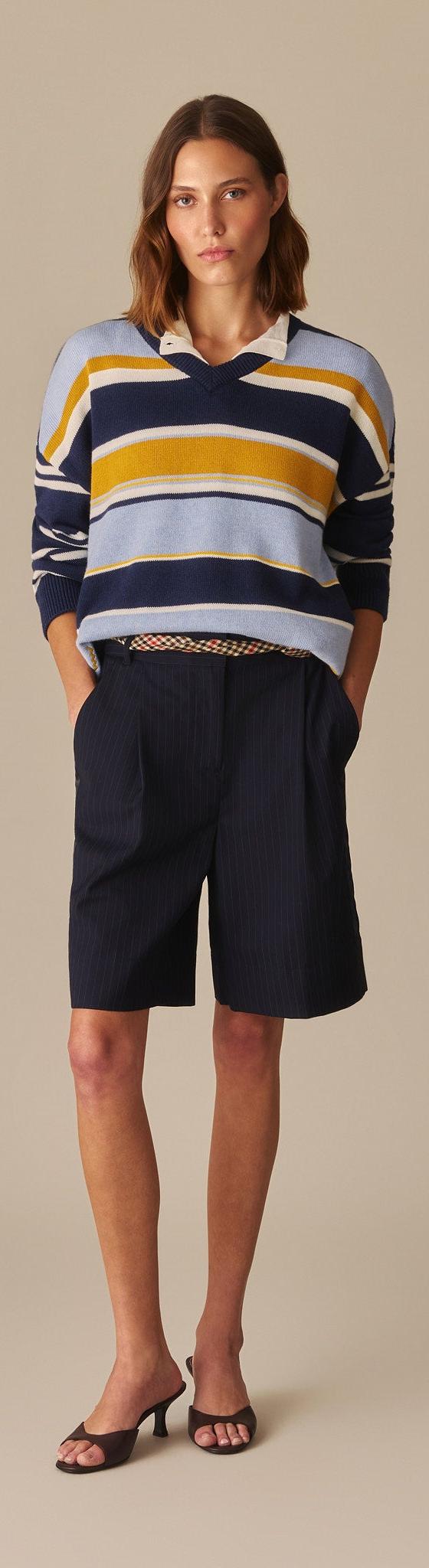 A woman wearing a navy, yellow and pale blue striped Rowing Blazers rugby-collar sweater with navy pleated shorts