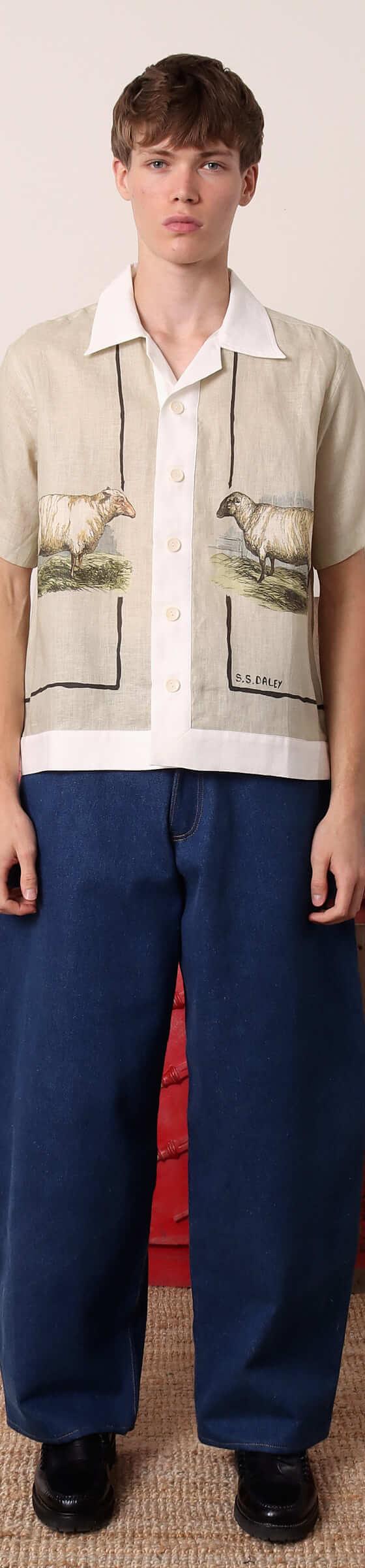 A man wearing an S.S.DALEY linen camp-collar shirt with pastoral sheep print and wide indigo jeans