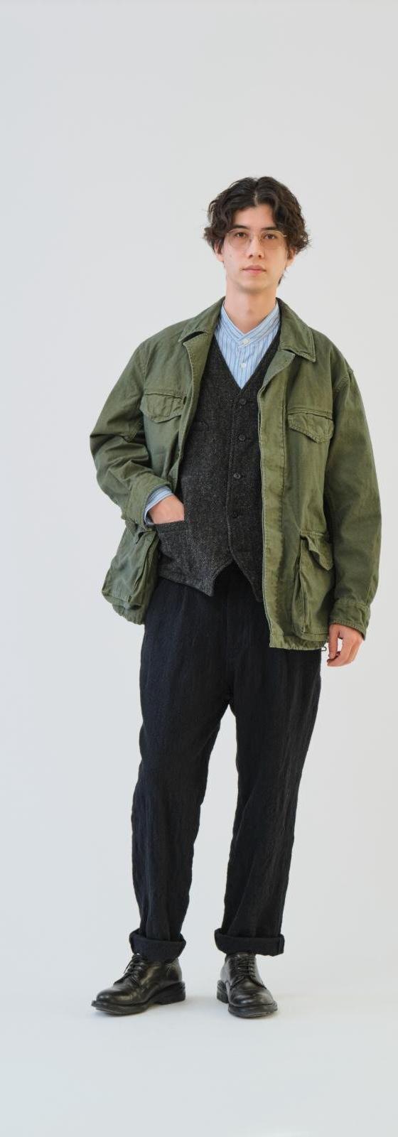 A man wearing an olive SAGE DE CRET field jacket over a knitted vest and black trousers