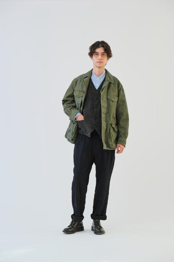 A man wearing an olive SAGE DE CRET field jacket over a knitted vest and black trousers