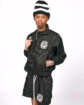 A man wearing a black Saint Vanity nylon zip jacket and matching cargo shorts with lion patches, and a striped beanie