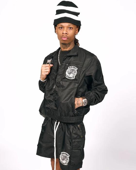 A man wearing a black Saint Vanity nylon zip jacket and matching cargo shorts with lion patches, and a striped beanie