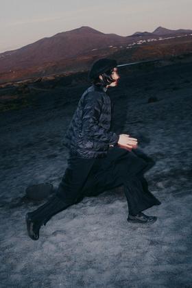 A man in a black quilted jacket, wide black trousers and Salomon Advanced trail sneakers striding across volcanic terrain at dusk