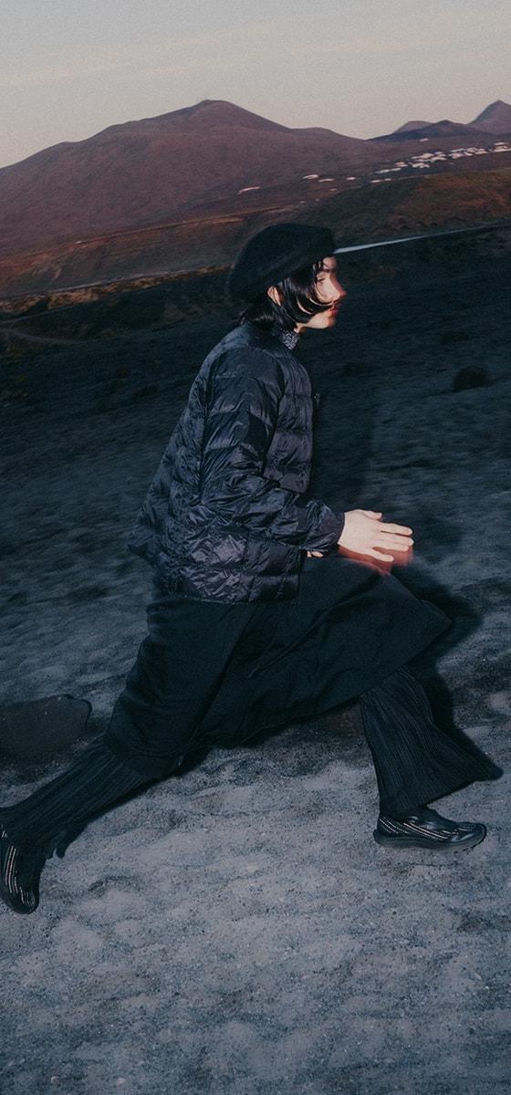 A man in a black quilted jacket, wide black trousers and Salomon Advanced trail sneakers striding across volcanic terrain at dusk
