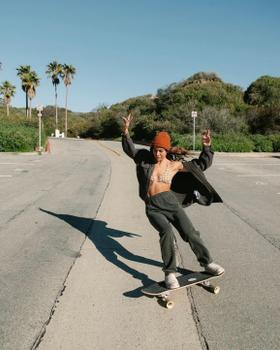 A woman wearing Sanuk slip-ons while skateboarding down a palm-lined coastal road