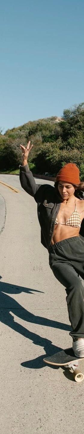 A woman wearing Sanuk slip-ons while skateboarding down a palm-lined coastal road