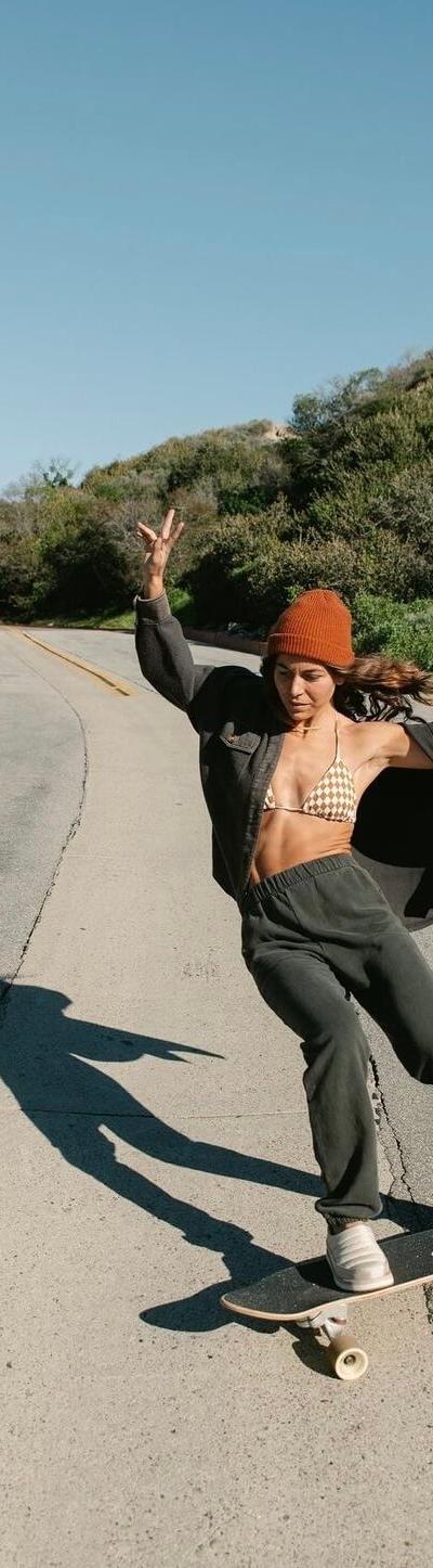 A woman wearing Sanuk slip-ons while skateboarding down a palm-lined coastal road