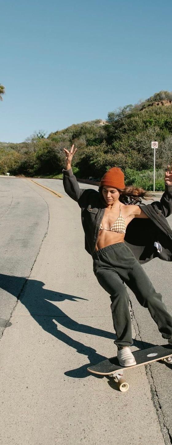 A woman wearing Sanuk slip-ons while skateboarding down a palm-lined coastal road