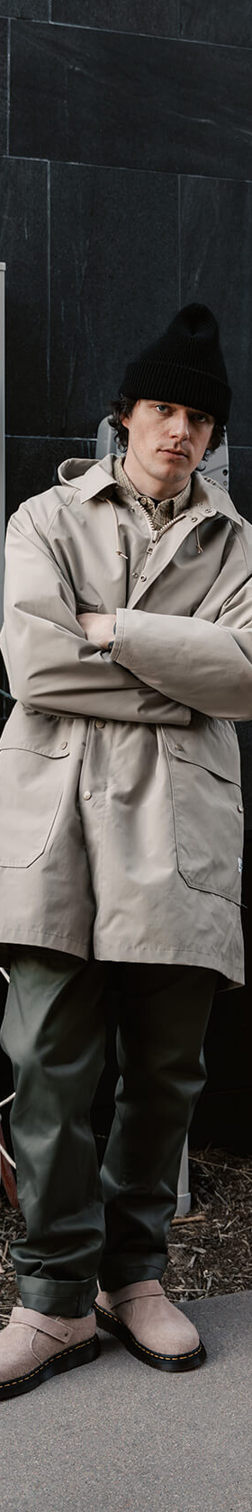 A man wearing a beige SASSAFRAS gardener coat with olive green trousers and a black beanie