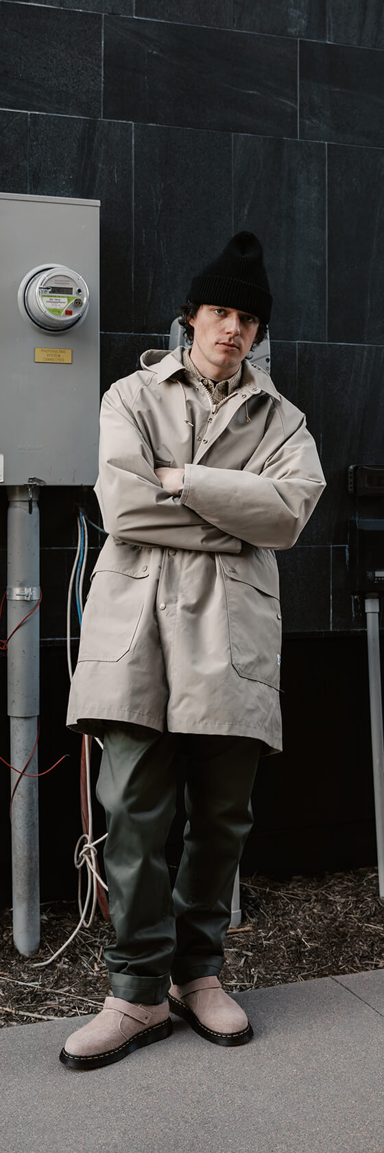 A man wearing a beige SASSAFRAS gardener coat with olive green trousers and a black beanie