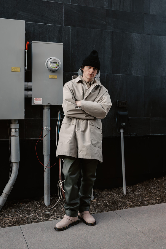 A man wearing a beige SASSAFRAS gardener coat with olive green trousers and a black beanie