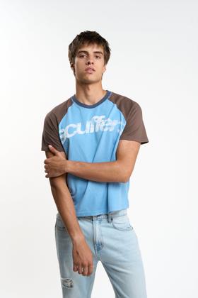 A man wearing a blue and brown raglan Scuffers logo T-shirt with light-wash jeans