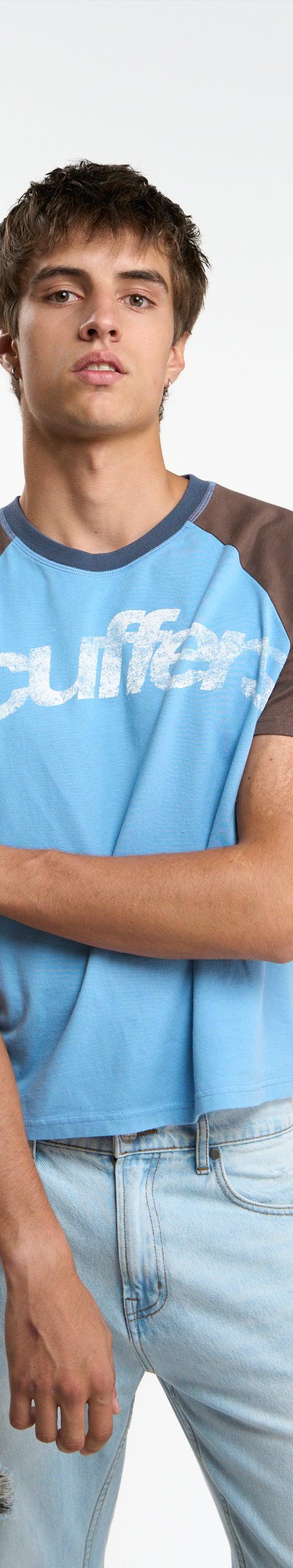 A man wearing a blue and brown raglan Scuffers logo T-shirt with light-wash jeans