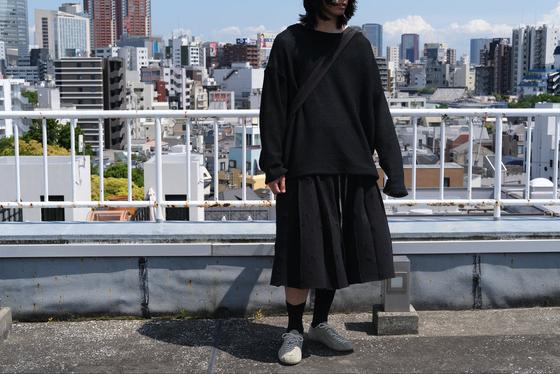 A man wearing a black SHINYAKOZUKA knit sweater and pleated skirt on a Tokyo rooftop