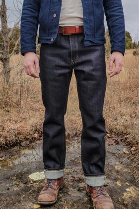 A man wearing raw selvedge denim jeans with cuffed hems, a dark denim chore jacket and brown leather boots