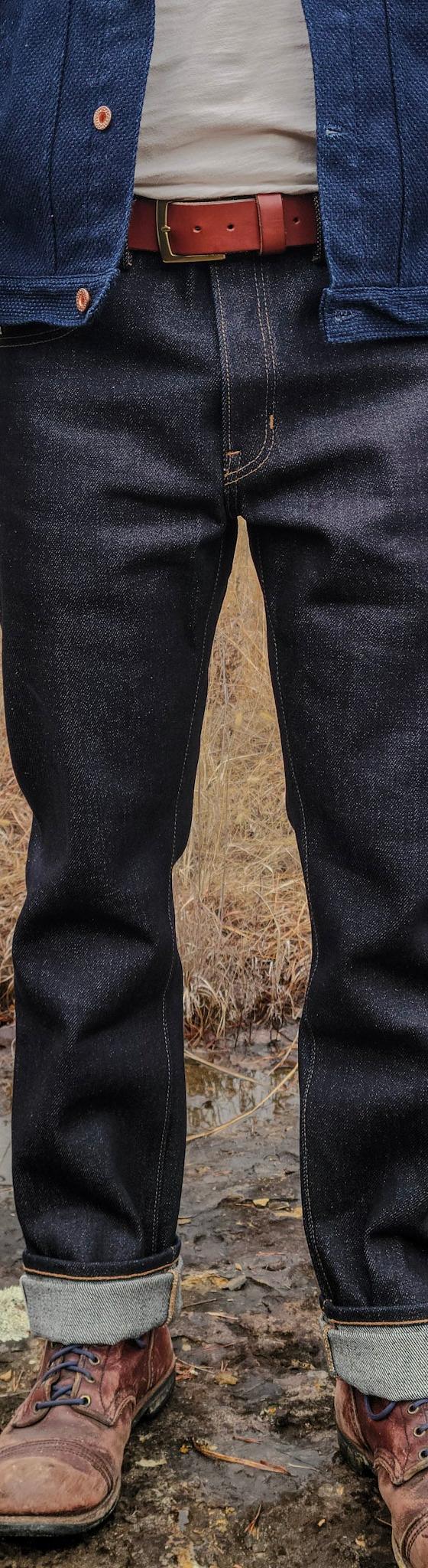 A man wearing raw selvedge denim jeans with cuffed hems, a dark denim chore jacket and brown leather boots