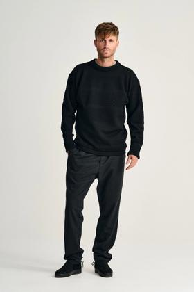 A man wearing a black S.N.S. Herning bobble-stitch Fisherman sweater with charcoal trousers
