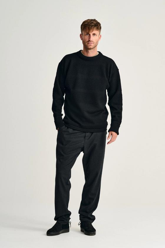 A man wearing a black S.N.S. Herning bobble-stitch Fisherman sweater with charcoal trousers