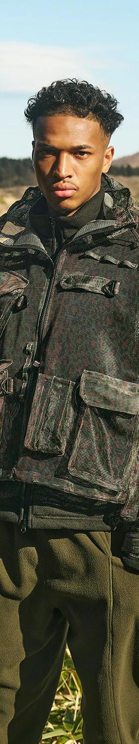 A man wearing a printed camouflage SOUTH2 WEST8 multi-pocket hunting jacket and olive trousers in an open marshland