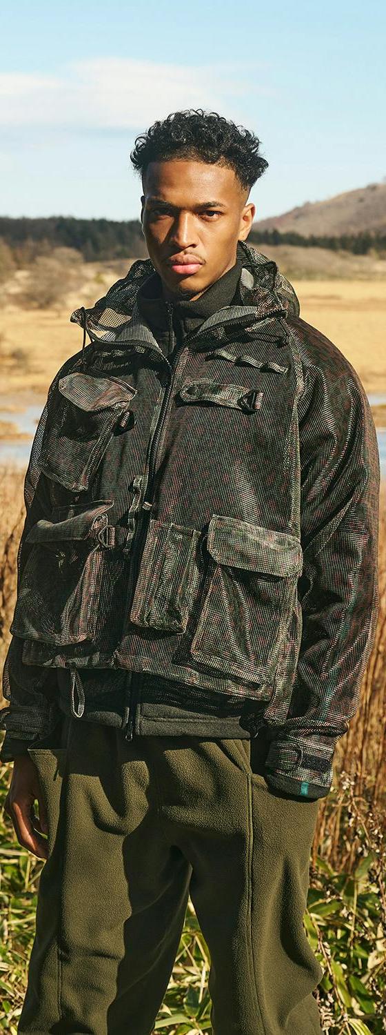 A man wearing a printed camouflage SOUTH2 WEST8 multi-pocket hunting jacket and olive trousers in an open marshland