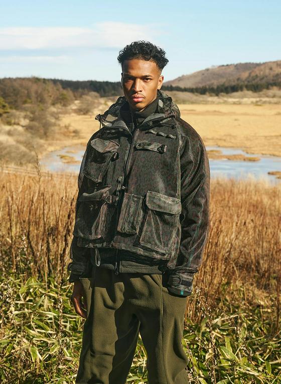 A man wearing a printed camouflage SOUTH2 WEST8 multi-pocket hunting jacket and olive trousers in an open marshland