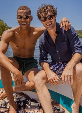 Two men on a boat, one wearing a navy linen shirt and printed swim shorts with sunglasses