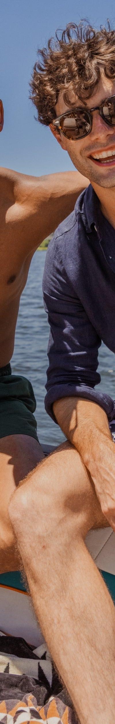 Two men on a boat, one wearing a navy linen shirt and printed swim shorts with sunglasses