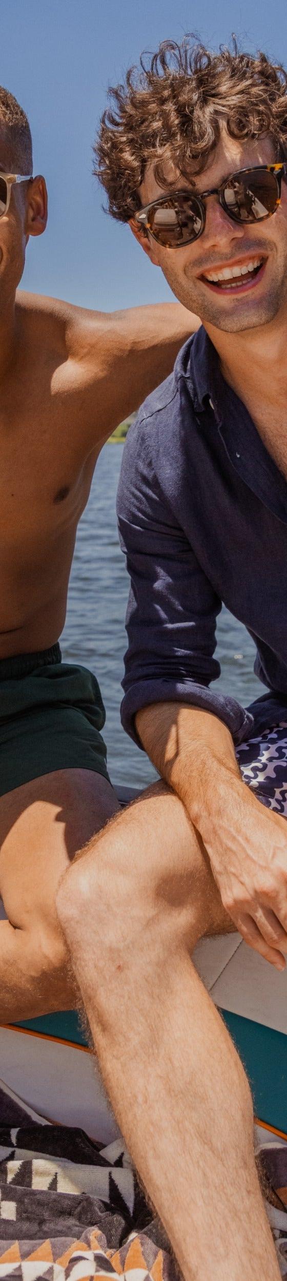 Two men on a boat, one wearing a navy linen shirt and printed swim shorts with sunglasses