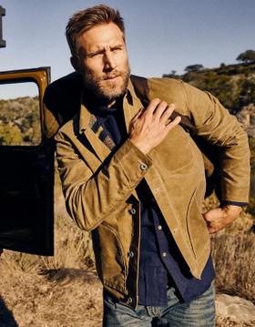 A bearded man wearing a tan waxed cotton jacket over a navy shirt and jeans beside a truck in the desert
