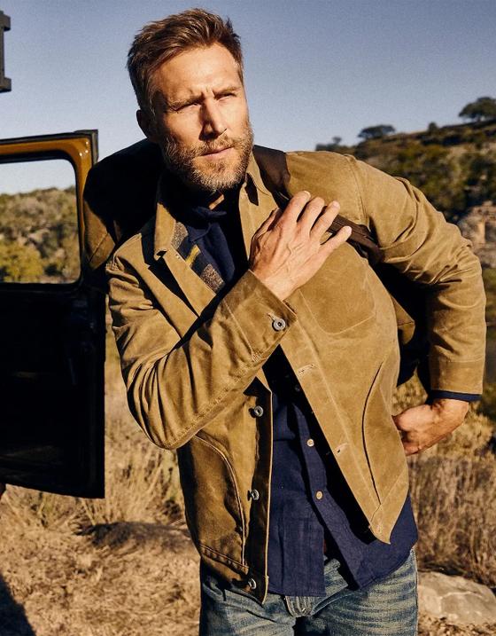 A bearded man wearing a tan waxed cotton jacket over a navy shirt and jeans beside a truck in the desert