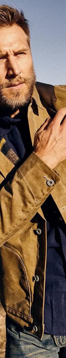 A bearded man wearing a tan waxed cotton jacket over a navy shirt and jeans beside a truck in the desert