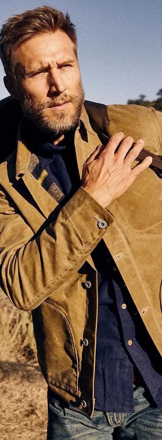 A bearded man wearing a tan waxed cotton jacket over a navy shirt and jeans beside a truck in the desert