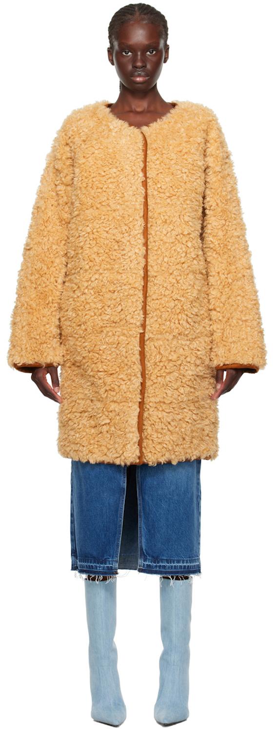 A woman wearing a beige faux fur Stand Studio coat with jeans and blue boots