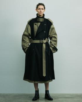 A person wearing a black and khaki deconstructed belted stein trench coat with black leather boots