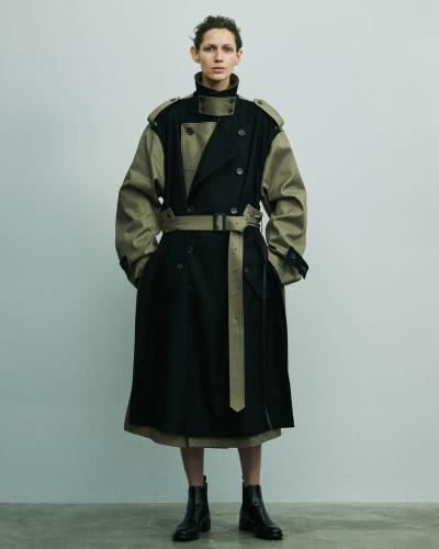 A person wearing a black and khaki deconstructed belted stein trench coat with black leather boots