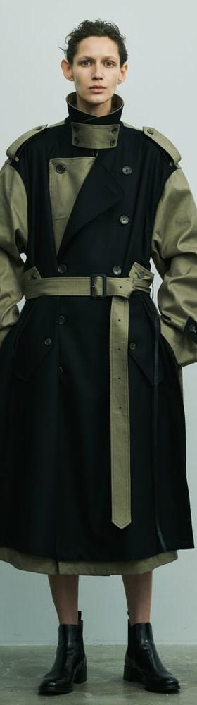 A person wearing a black and khaki deconstructed belted stein trench coat with black leather boots