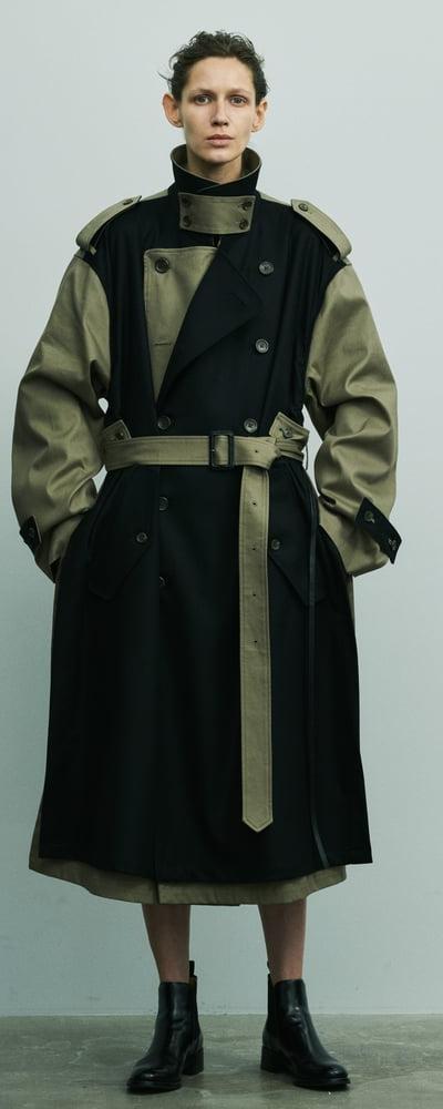 A person wearing a black and khaki deconstructed belted stein trench coat with black leather boots