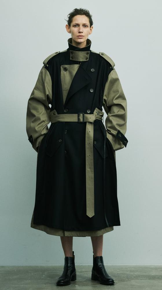A person wearing a black and khaki deconstructed belted stein trench coat with black leather boots