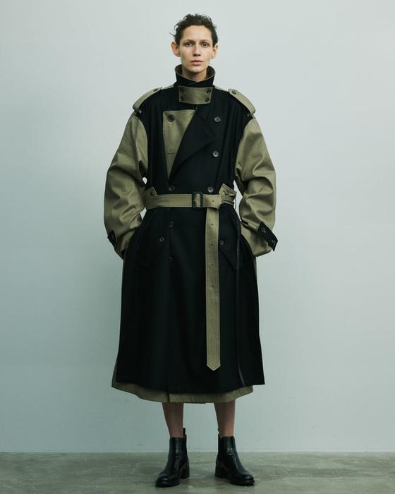 A person wearing a black and khaki deconstructed belted ssstein trench coat with black leather boots
