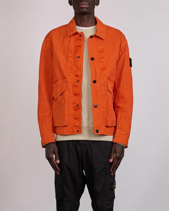 A man wearing an orange Stone Island nylon overshirt jacket with the compass badge, a beige crewneck and black cargo trousers