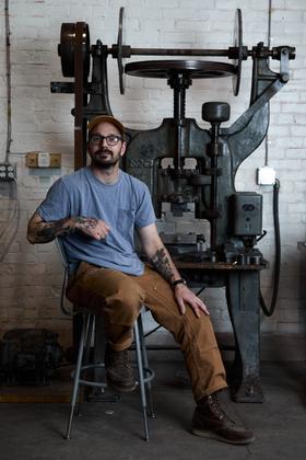 A man wearing a grey tee, brown workwear trousers and a Studebaker Metals cuff, seated beside a drop hammer in a brick workshop