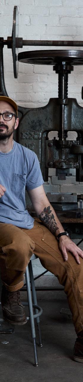 A man wearing a grey tee, brown workwear trousers and a Studebaker Metals cuff, seated beside a drop hammer in a brick workshop