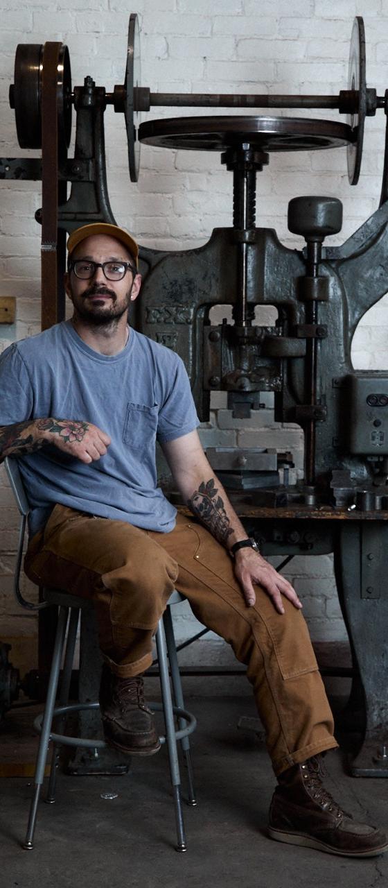 A man wearing a grey tee, brown workwear trousers and a Studebaker Metals cuff, seated beside a drop hammer in a brick workshop