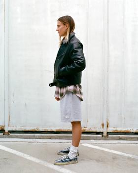 A person wearing a black leather jacket over a faded plaid shirt with grey Stüssy sweat shorts and sneakers