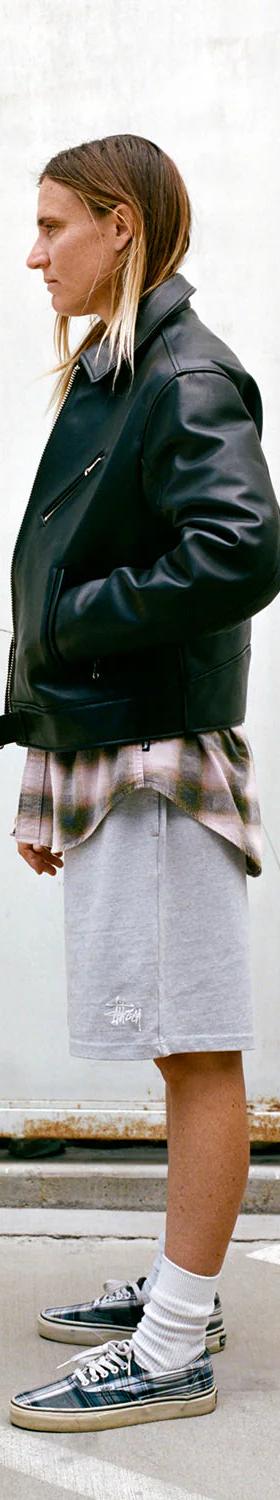 A person wearing a black leather jacket over a faded plaid shirt with grey Stüssy sweat shorts and sneakers