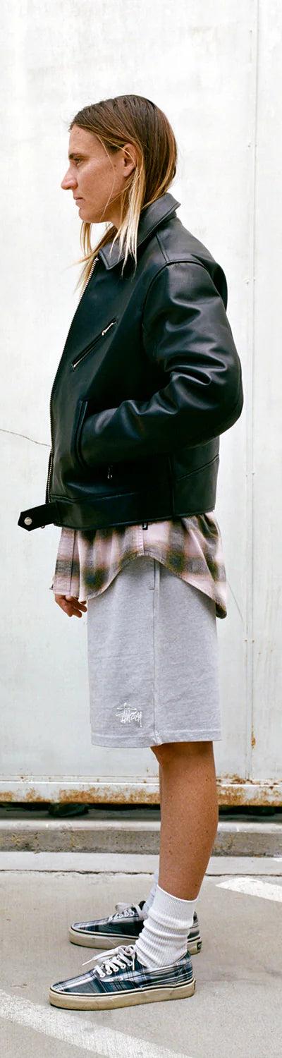A person wearing a black leather jacket over a faded plaid shirt with grey Stüssy sweat shorts and sneakers